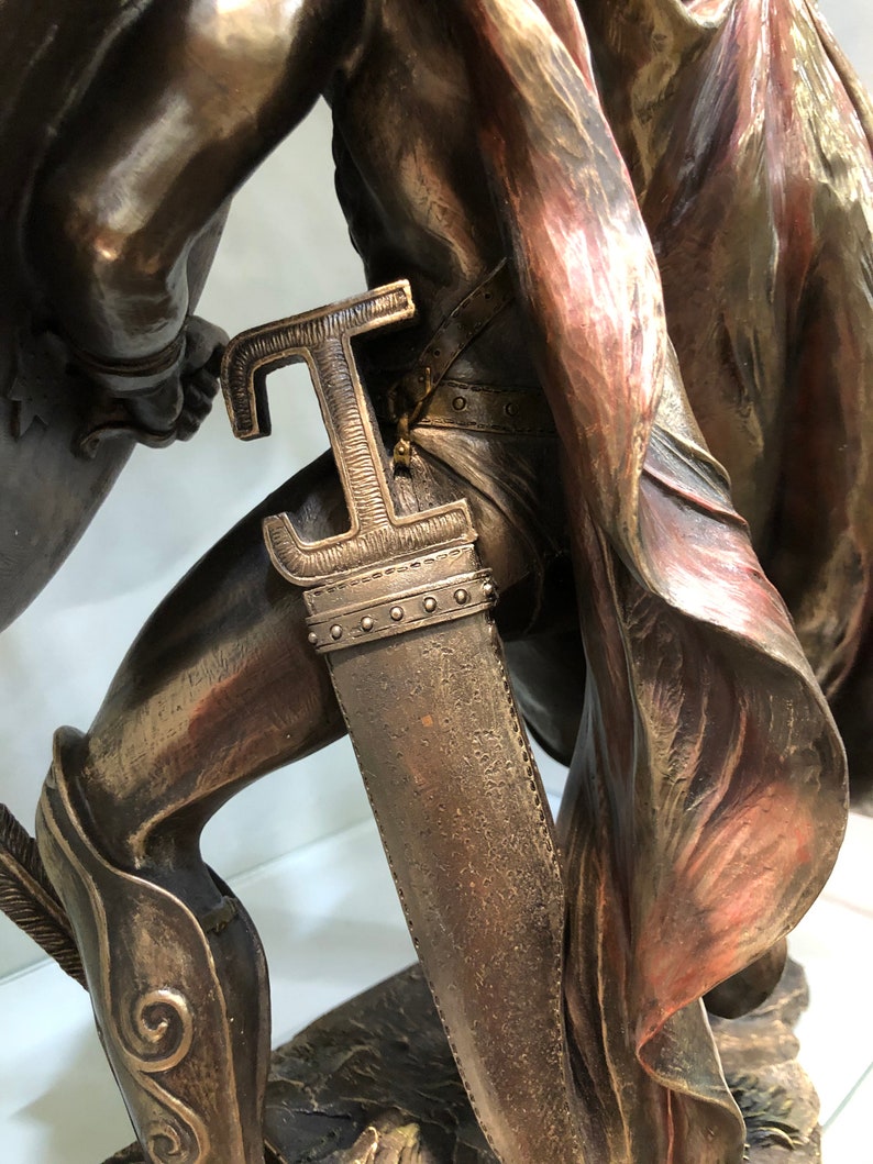 May include: Bronze statue of a warrior holding a sword and shield. The warrior is wearing a red cape and has a detailed, textured surface.