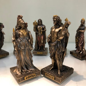 May include: Bronze figurines of Athena and Apollo, Greek gods, standing on bases with their names inscribed. The figurines are detailed and have a golden finish.