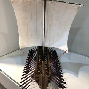 Puede incluir: Un modelo detallado de un barco trirreme griego antiguo de madera con una vela blanca y remos morados. El barco est&aacute; en exhibici&oacute;n en un estante de vidrio.