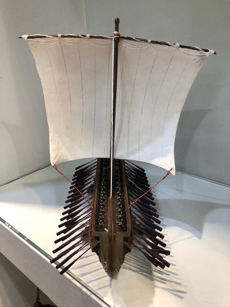 Puede incluir: Un modelo detallado de un barco trirreme griego antiguo de madera con una vela blanca y remos morados. El barco est&aacute; en exhibici&oacute;n en un estante de vidrio.