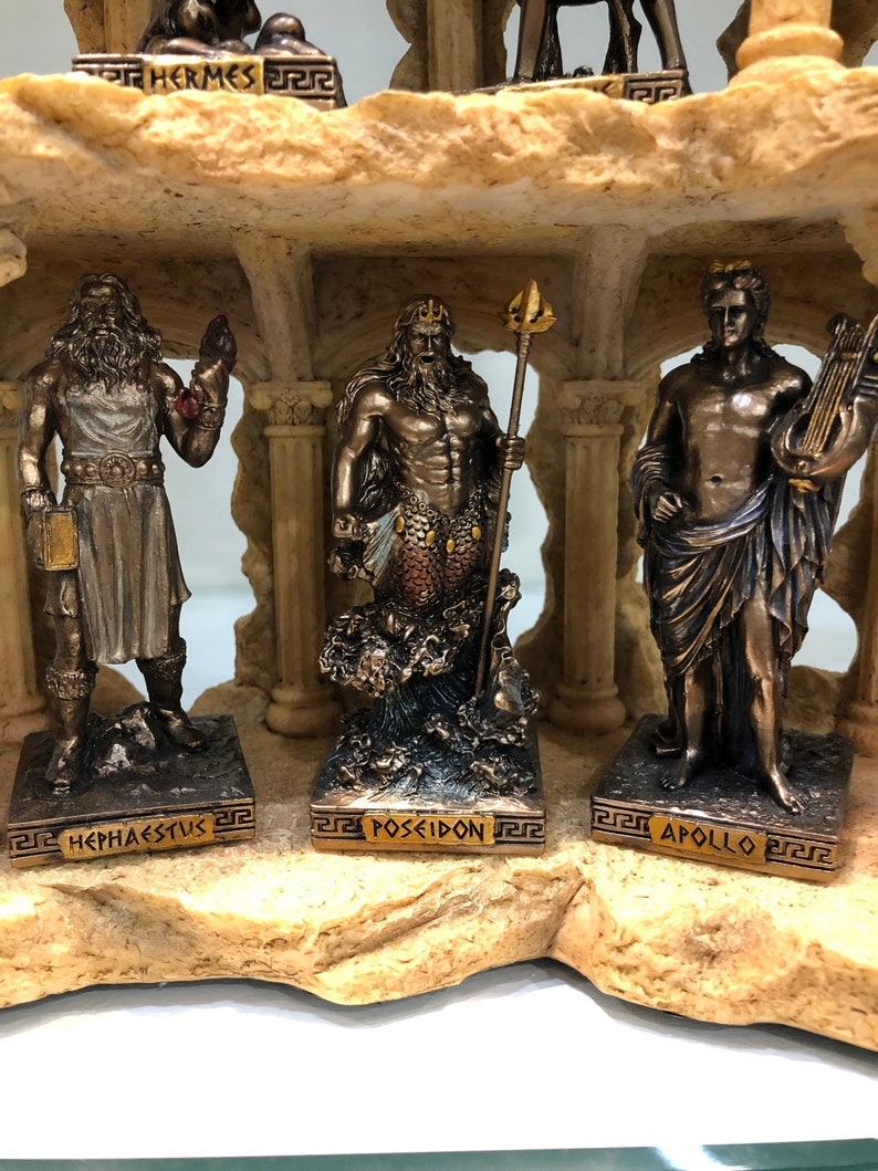 May include: A set of six bronze Greek god figurines, each standing on a separate base with their name inscribed in gold lettering. The figurines are arranged on a rocky, beige-colored base. The gods depicted are Hermes, Hephaestus, Poseidon, Apollo, Zeus, and Athena.