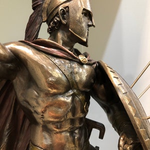 May include: Bronze statue of a Spartan warrior wearing a helmet with a red plume, a red cape, and holding a shield and a sword. The warrior is depicted in a fighting pose.