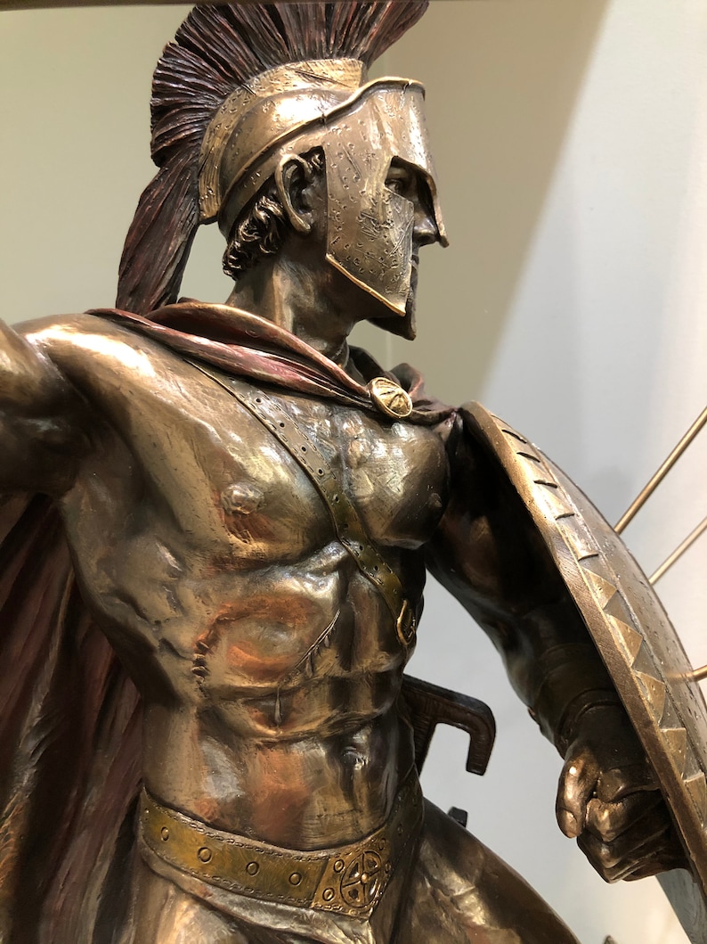 May include: Bronze statue of a Spartan warrior wearing a helmet with a red plume, a red cape, and holding a shield and a sword. The warrior is depicted in a fighting pose.