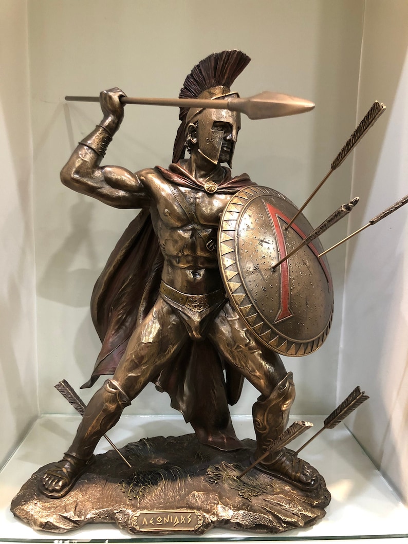 May include: Bronze statue of a Spartan warrior holding a spear and shield. The warrior is wearing a helmet with a red plume and a red cape. The shield has a red triangle on it. There are arrows sticking out of the shield and the ground. The base of the statue is inscribed with the word "LEONIDAS".