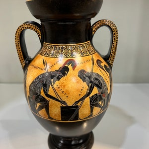 May include: Black and orange Greek amphora with two handles and a black base. The amphora is decorated with a black and white image of two warriors with spears, and the text "AIA".