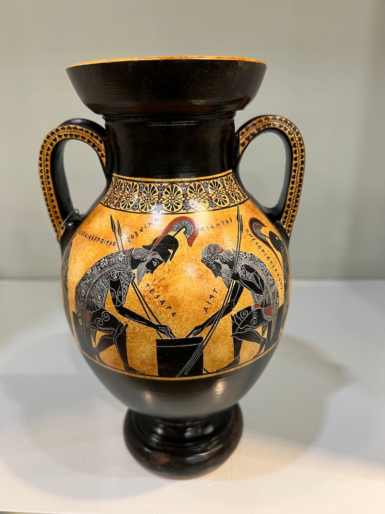 May include: Black and orange Greek amphora with two handles and a black base. The amphora is decorated with a black and white image of two warriors with spears, and the text "AIA".