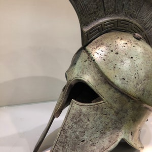 May include: A bronze Greek warrior helmet with a plume on top and a decorative band around the crown. The helmet has a face mask and a chin strap.