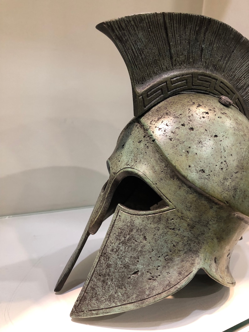 May include: A bronze Greek warrior helmet with a plume on top and a decorative band around the crown. The helmet has a face mask and a chin strap.