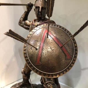 May include: Bronze sculpture of a Spartan warrior holding a spear and shield. The shield is decorated with a red triangle and the warrior is wearing a helmet with a red plume.