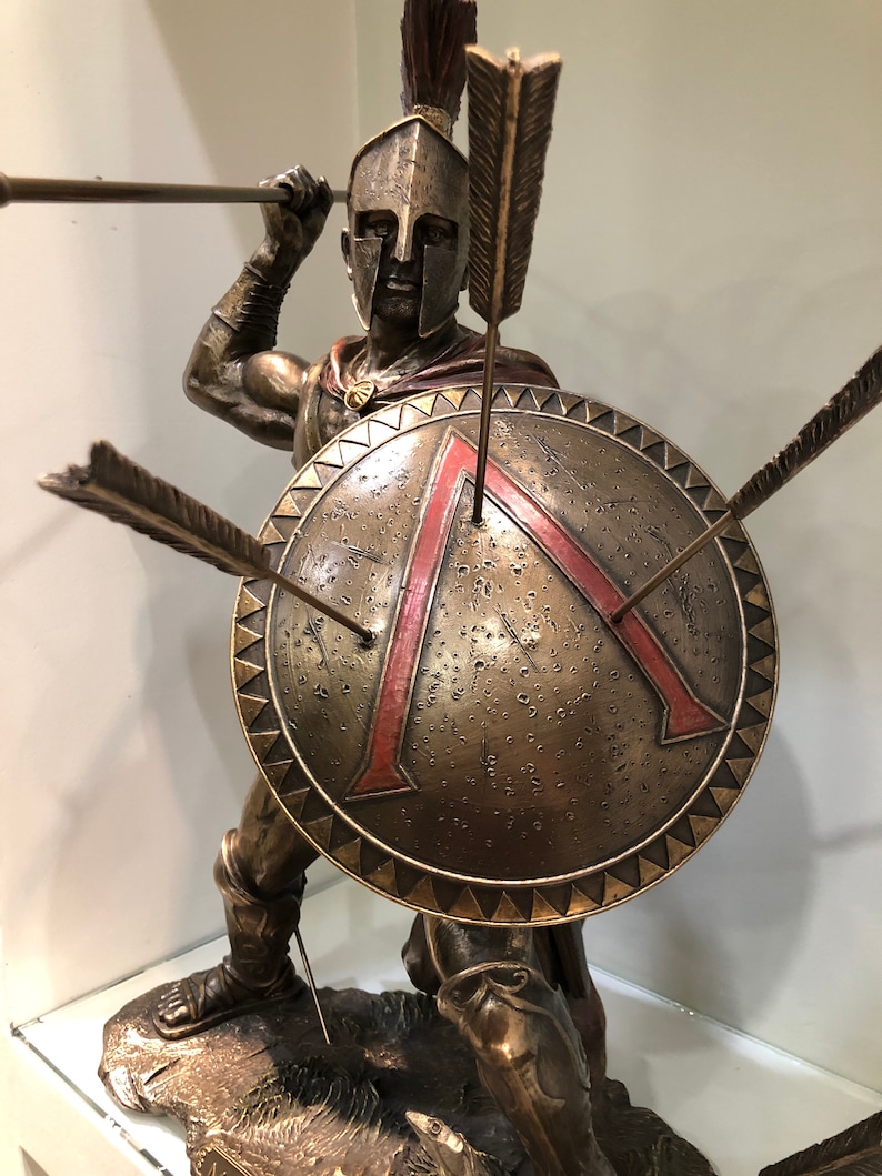 May include: Bronze sculpture of a Spartan warrior holding a spear and shield. The shield is decorated with a red triangle and the warrior is wearing a helmet with a red plume.