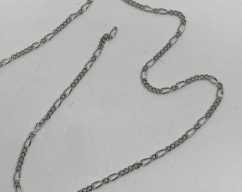 Könnte beinhalten: Eine silberne Kettenkette mit einem zarten Figaro-Gliederdesign. Die Kette liegt flach auf einer weißen Oberfläche.