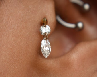 May include: A close-up of a person's ear with two earrings. The top earring is a gold dangle earring with two clear stones. The bottom earring is a gold stud earring with a purple stone.