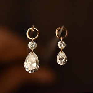 Op de afbeelding: Een paar gouden oorbellen met een klassiek ontwerp. Elke oorbel heeft een kleine, ronde diamant boven een grotere, peervormige diamant. De oorbellen zijn opgehangen aan een gouden ring.