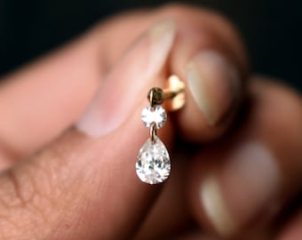 May include: A gold-toned stud earring featuring a small, round, clear faceted stone connected to a larger, pear-shaped, clear faceted drop stone. The earring has a thin metal post and a flat back component, showcasing a delicate and refined design.