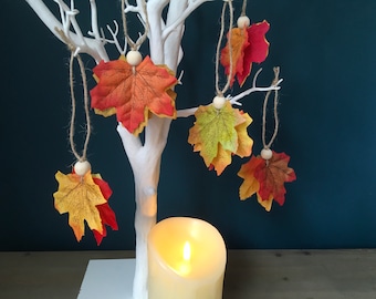 Può includere: Un albero di rami bianchi con cinque ornamenti a forma di foglie d'acero di colore autunnale appesi. Le foglie sono rosse, arancioni e gialle. Una candela bianca è davanti all'albero.