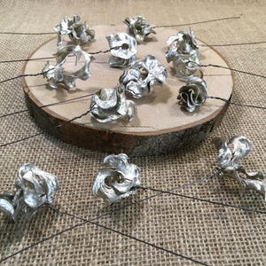 May include: Silver metal flower decorations with black wire stems are displayed on a wooden slice and a burlap background. The flowers have different designs, some open and others tightly wound.
