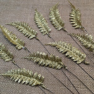 May include: Ten gold glitter fern sprigs with wire stems. The ferns are arranged on a hessian background.