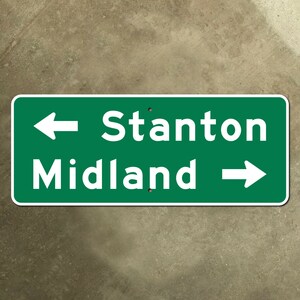 Puede incluir: Señal de carretera verde con texto blanco y flechas. La señal indica las direcciones a Stanton y Midland. Una flecha que apunta hacia la izquierda indica Stanton y una flecha que apunta hacia la derecha indica Midland.