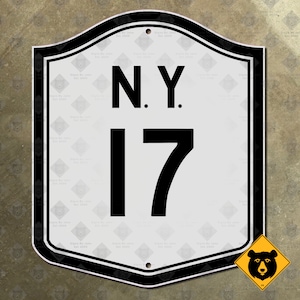 Op de afbeelding: Een wit en zwart verkeersbord met de tekst "N.Y. 17". Het bord heeft een zwarte rand en een klein waarschuwingsbord voor beren in de hoek. Gemaakt door "Signs By Jake Est. 2005".
