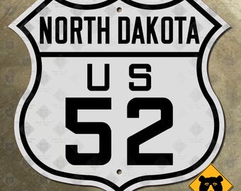 Könnte beinhalten: Ein weißes Metallschild mit schwarzer Beschriftung. Das Schild ist schildförmig und trägt oben den Text "NORTH DAKOTA" und in der Mitte "US 52".