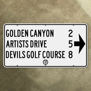 May include: White sign with black text and an arrow pointing right. The sign lists directions to Golden Canyon (2 miles), Artists Drive (5 miles), and Devils Golf Course (8 miles).
