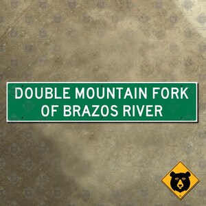 Könnte beinhalten: Ein grünes rechteckiges Schild mit weißem Text, der "DOUBLE MOUNTAIN FORK OF BRAZOS RIVER" lautet. Ein gelbes rautenförmiges Schild mit einer schwarzen Bärgrafik befindet sich unten rechts.
