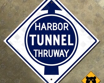 May include: A white and blue diamond-shaped sign with the words "HARBOR TUNNEL THRUWAY" in white text inside a blue circle. An upward-pointing arrow is at the top. A small yellow sign with a black bear is in the lower right corner.