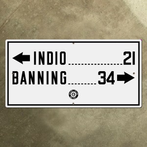 May include: White sign with black text and arrows indicating directions to Indio (21 miles) and Banning (34 miles). The sign also features a black and white logo for the "Auto Club of So. Cal."