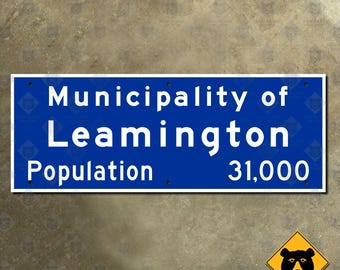 May include: A rectangular blue sign with white text that reads "Municipality of Leamington Population 31,000." The sign is mounted on a textured surface. A yellow warning sign with a black bear graphic is in the lower right corner.