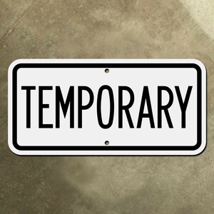 Może przedstawiać: Prostokątny biały znak z napisem "TEMPORARY" pogrubioną czarną czcionką. Znak ma czarną obwódkę i dwa wstępnie wywiercone otwory do montażu. Tło to teksturowana powierzchnia o neutralnym odcieniu.