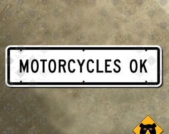 Könnte beinhalten: Ein rechteckiges weißes Metallschild mit schwarzem Rand und dem schwarzen Text "MOTORCYCLES OK" in der Mitte. Das Schild verfügt über vier kleine kreisförmige Befestigungslöcher, die sich jeweils in der Nähe der Ecken befinden.