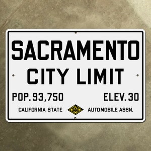 May include: White rectangular sign with black text that reads "Sacramento City Limit". Below the text is "Pop. 93,750 California State" and "Elev. 30 Automobile Assn.". A yellow diamond with black text that reads "California State Automobile Association" is in the lower right corner.