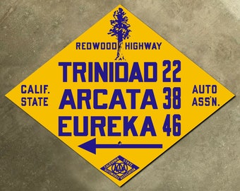 May include: A yellow diamond-shaped sign with blue text and a blue tree silhouette. The sign reads "Redwood Highway", "Trinidad 22", "Arcata 38", "Eureka 46", "Calif. State", and "Auto Assn."