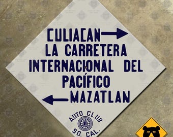Op de afbeelding: Een ruitvormig bord met donkerblauwe tekst met aanwijzingen naar Culiacan en Mazatlan op de Carretera Internacional del Pacifico. Het bord heeft ook het logo van Auto Club So. Cal. en een klein geel berenbord.