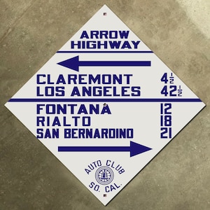 May include: A white diamond-shaped sign with blue text and arrows. The sign reads "Arrow Highway" and lists distances to Claremont, Los Angeles, Fontana, Rialto, and San Bernardino. The sign also includes the logo for the "Auto Club So. Cal."