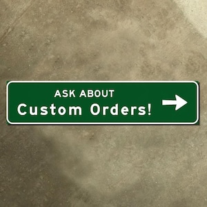 May include: A green rectangular sign with white text that reads "ASK ABOUT Custom Orders!" and a white arrow pointing right. The sign has a white border and is set against a textured, neutral-toned background.