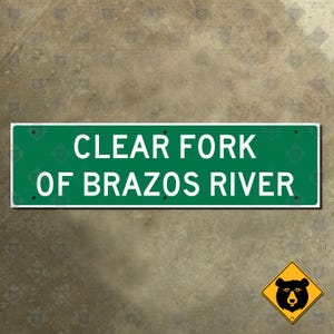 Könnte beinhalten: Ein rechteckiges grünes Schild mit weißem Text "CLEAR FORK OF BRAZOS RIVER". Das Schild hat einen weißen Rand und ist mit schwarzen Schrauben befestigt. Ein gelbes Diamantschild mit einer schwarzen Bärengrafik befindet sich unten rechts.