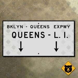 May include: A rectangular white metal road sign with a black border and black text reading BKLYN - QUEENS EXPWY and QUEENS - L.I. with two downward-pointing black arrows. The sign features two small circular mounting holes at the top and bottom center.