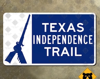 Op de afbeelding: Een rechthoekig blauw en wit bord met de tekst "TEXAS INDEPENDENCE TRAIL" in het wit. Een silhouet van een geweer staat aan de linkerkant. Een geel diamantbord met een zwarte beer staat in de rechter benedenhoek.