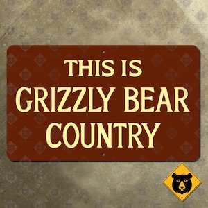 May include: A rectangular brown sign with the text "THIS IS GRIZZLY BEAR COUNTRY" in large, yellow capital letters. A small yellow sign with a black bear graphic is in the lower right corner. The sign has two holes for mounting.
