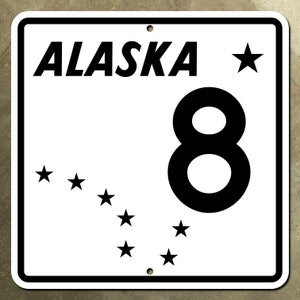 May include: White sign with black text and a black number 8. The sign says "ALASKA" and has a black star in the top right corner. There are seven black stars in the bottom left corner, forming a constellation.