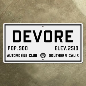 Puede incluir: Señal de metal blanco con texto negro que dice "DEVORE", "POP. 900 AUTOMOBILE CLUB", y "ELEV. 2510 SOUTHERN CALIF.". Un logotipo en blanco y negro de una rueda de automóvil con las palabras "AUTO CLUB SO. CAL." está en el centro del letrero.