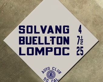May include: A white diamond-shaped sign with blue text. The sign reads "Solvang 4, Buellton 7 1/2, Lompoc 25". The sign also has a blue circle with the text "Auto Club So. Cal."