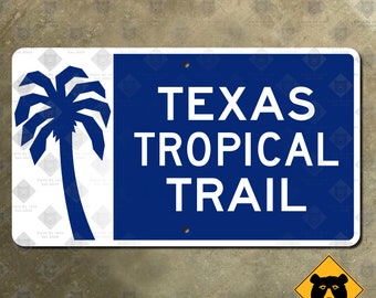 Op de afbeelding: Een rechthoekig bord met een witte achtergrond en een blauw gedeelte met de tekst "TEXAS TROPICAL TRAIL". Een blauwe palmboomafbeelding staat links. Het bord heeft twee gaten om te monteren.