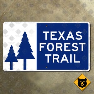 May include: A rectangular sign with a white background and a blue section. The blue section has the words "TEXAS FOREST TRAIL" in white. Two blue pine tree silhouettes are on the left. A yellow bear sign is in the bottom right.