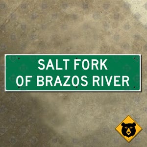 Könnte beinhalten: Ein rechteckiges grünes Schild mit weißem Text "SALT FORK OF BRAZOS RIVER". Das Schild hat einen weißen Rand und ist auf einer strukturierten Oberfläche montiert. Ein gelbes rautenförmiges Schild mit einer schwarzen Bärgrafik befindet sich unten rechts.