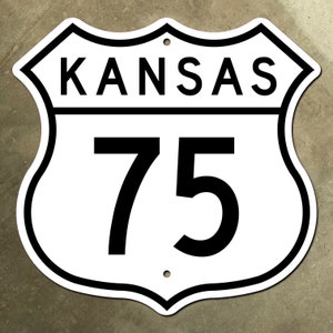 Puede incluir: Señal de carretera blanca y negra con el texto "KANSAS" y el número "75" en negro.