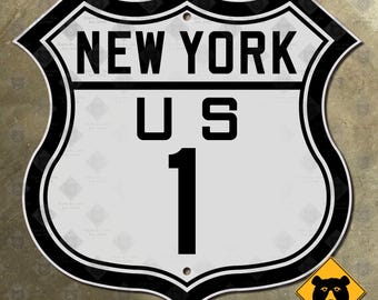 Könnte beinhalten: Ein schildförmiges Metall-Straßenschild mit weißem Hintergrund und schwarzem Rand. Der Text NEW YORK ist oben in Schwarz aufgedruckt, gefolgt von U S und der Zahl 1 darunter. Es gibt zwei kleine kreisförmige Löcher zur Befestigung.