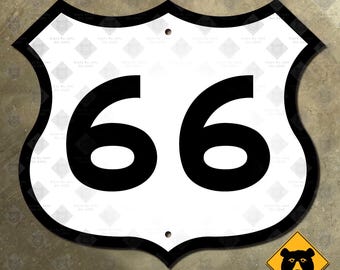 Op de afbeelding: Een wit metalen bord met zwarte letters, in de vorm van een schild, met het nummer "66" erop. Het bord is een replica van een klassiek US Route 66 wegenbord.