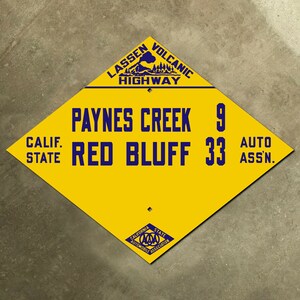 Pode incluir: Um sinal amarelo em forma de diamante com texto azul e um logotipo azul. O sinal diz "Lassen Volcanic Highway", "Paynes Creek 9", "Calif. State Red Bluff 33", e "Auto Assn.". O logotipo é uma imagem estilizada de um vulcão com árvores e o texto "California State Automobile Association".